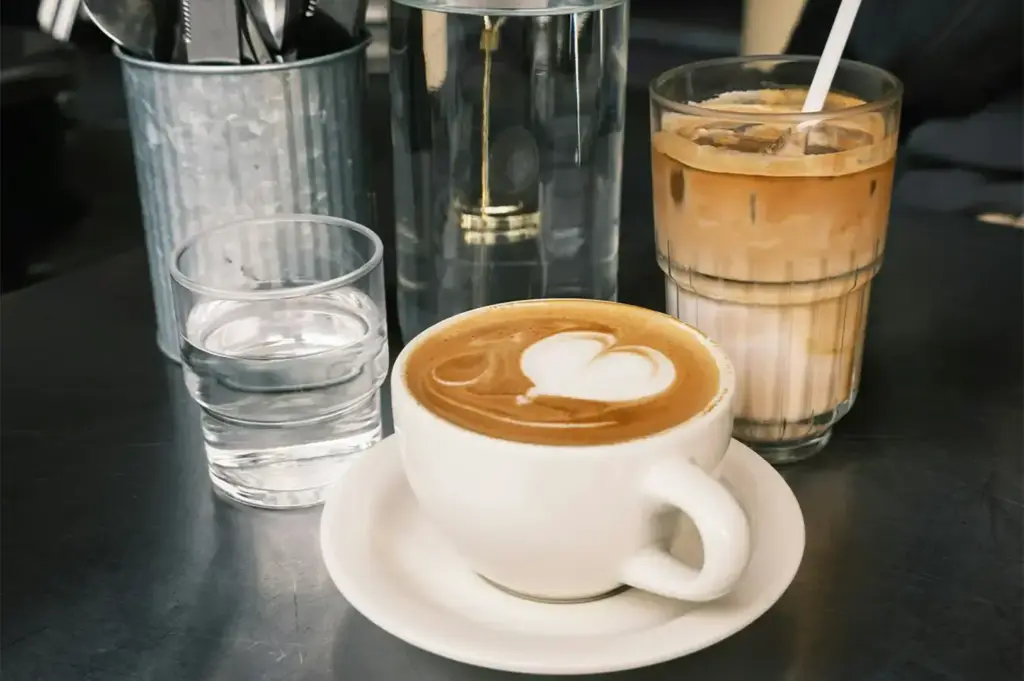 Close-up of a creamy cappuccino with heart-shaped latte art beside an iced latte, glass of water, and metal utensil holder on a dark café countertop. The warm tones and coffee presentation highlight a stylish specialty coffee shop environment.
