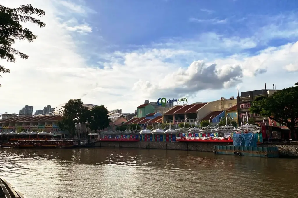 Scenic view of the Singapore River at Clarke Quay, featuring colorful colonial shop warehouses, the iconic CQ Clarke Quay sign, and river cruise boats under a blue sky.