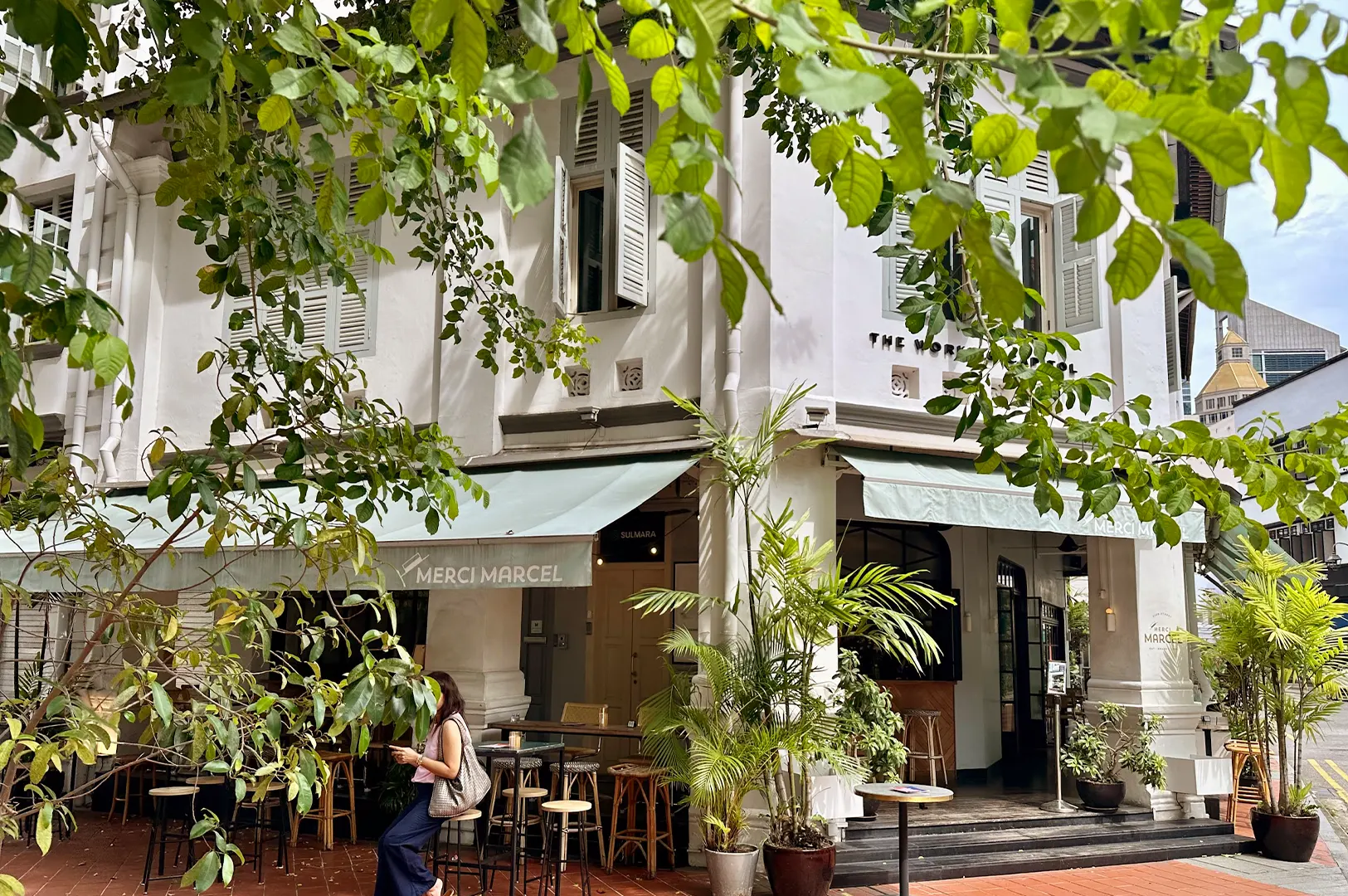 Exterior view of Merci Marcel on Club Street, featuring its elegant white shophouse architecture, lush greenery, and inviting outdoor terrace seating.