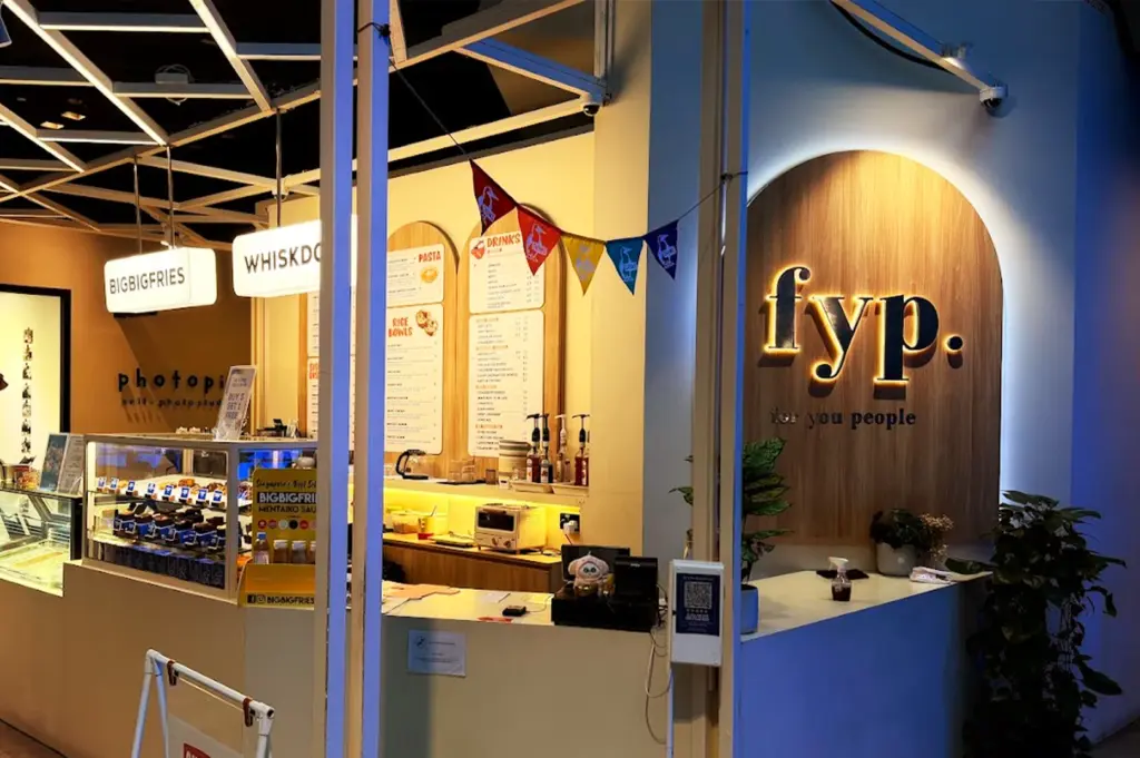 The brightly lit, trendy facade of FYP Cafe in Singapore, featuring its signature illuminated sign, and menu board listing pasta dishes, creating an inviting and comfortable atmosphere.