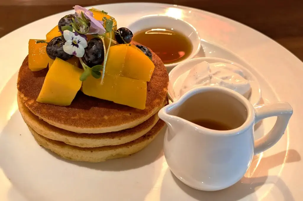 A close-up of a delicious Wild Honey breakfast dish featuring fluffy pancakes topped with fresh mango, blueberries, edible flowers, syrup, and whipped cream.