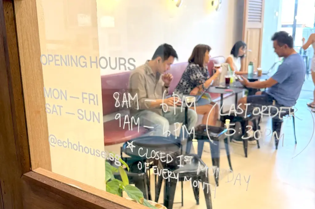 A view through the glass door of Echo Cafe, displaying its weekly opening hours and the cozy, calm interior where patrons sit at small tables to enjoy their coffee.