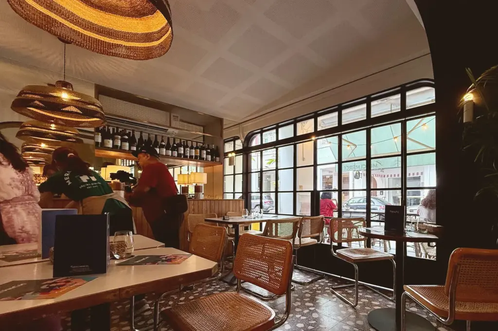 The warm, sunlit interior of Merci Marcel Club Street, highlighting rattan bistro chairs, classic mosaic tile flooring, and a relaxed French café atmosphere.