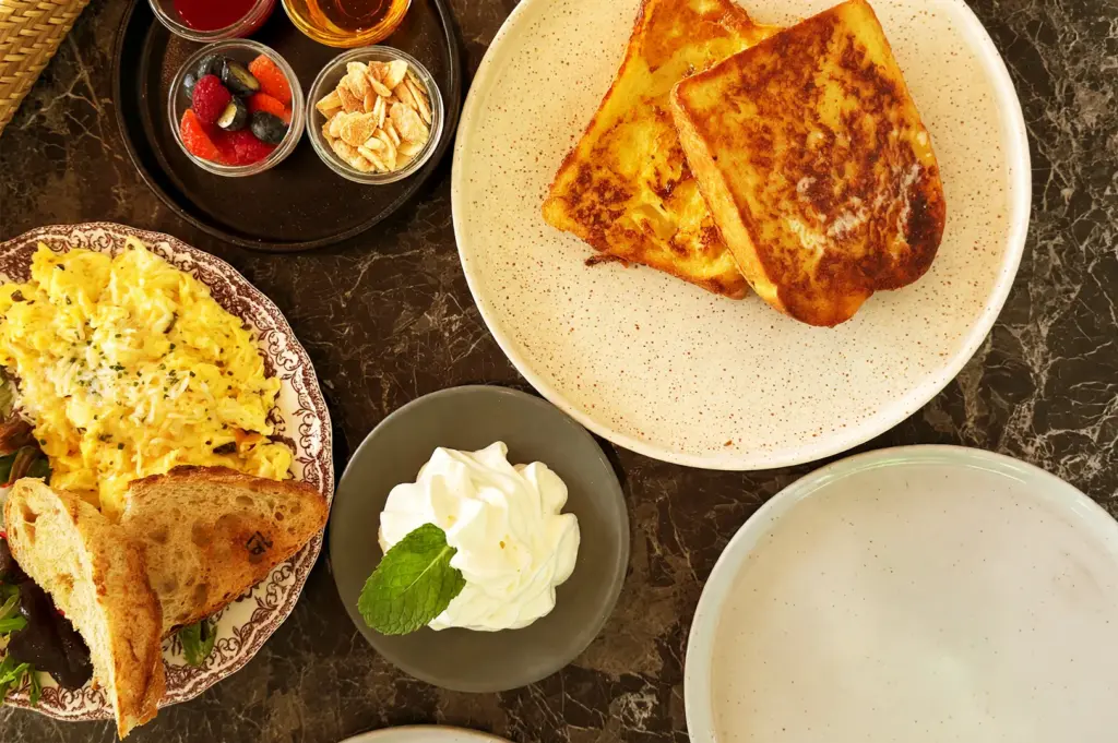 A gourmet brunch spread at Merci Marcel featuring scrambled eggs on sourdough toast, golden French toast, a bowl of fresh berries, and a side of whipped cream on a marble tabletop.