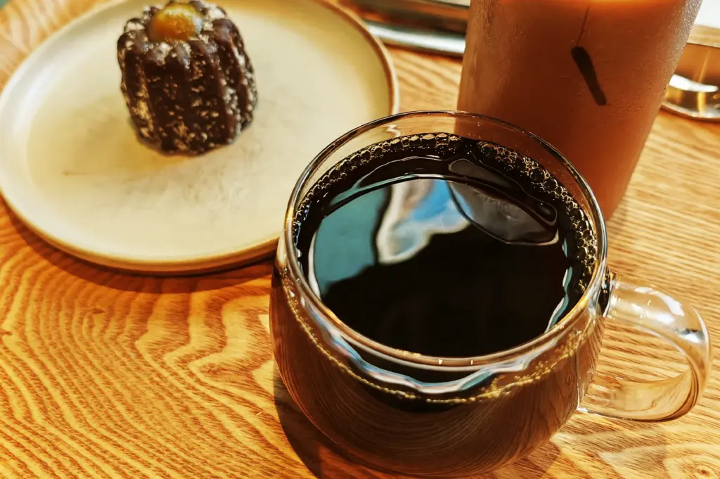 A refined coffee experience at Blue Bottle Coffee, displaying a clear glass cup of black coffee paired with a decadent canelé pastry on a wooden table.