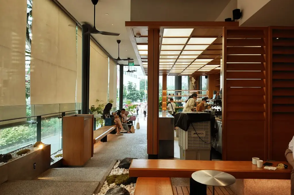 A sleek, modern interior of an Alchemist cafe branch, featuring minimalist wooden bench seating, bright overhead lighting, and stone accents.