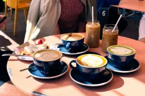 A round cafe table in Singapore filled with colorful cups of latte art coffee, iced beverages, and a sweet pastry.