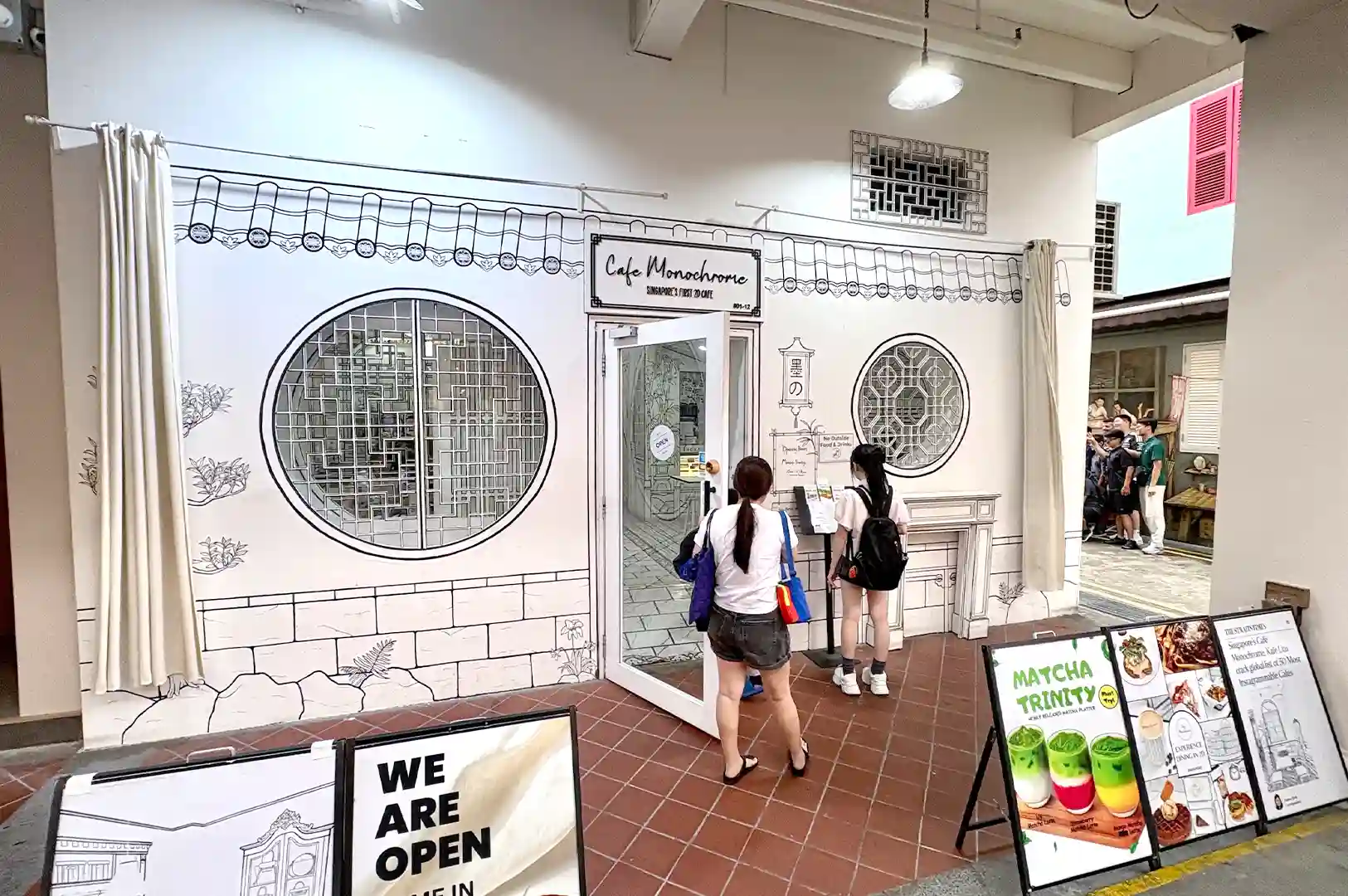 The front entrance and exterior facade of Cafe Monochrome in Singapore, showcasing unique 2D comic-style hand-drawn black and white illustrations.