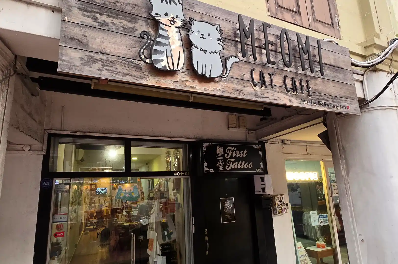Wooden storefront sign and entrance of Meomi Cat Cafe in Singapore, featuring painted cartoon cat illustrations and welcoming visitors.