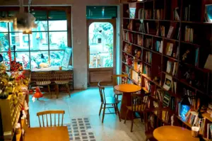 Cozy cafe interior with wooden tables and chairs, lined with bookshelves. Soft lighting creates a warm, inviting atmosphere. Large windows show lush plants outside.