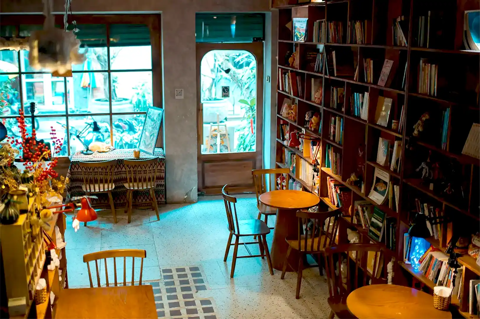 Cozy cafe interior with wooden tables and chairs, lined with bookshelves. Soft lighting creates a warm, inviting atmosphere. Large windows show lush plants outside.
