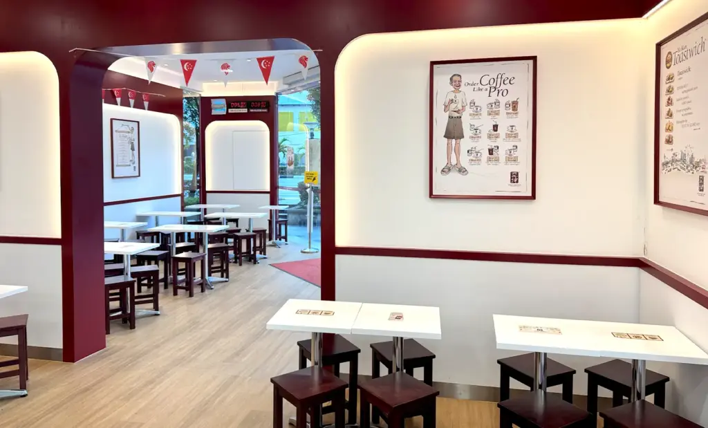 Interior view of a modern-traditional Ya Kun Kaya Toast cafe featuring white tables, dark wooden stools, and instructional posters on ordering coffee.