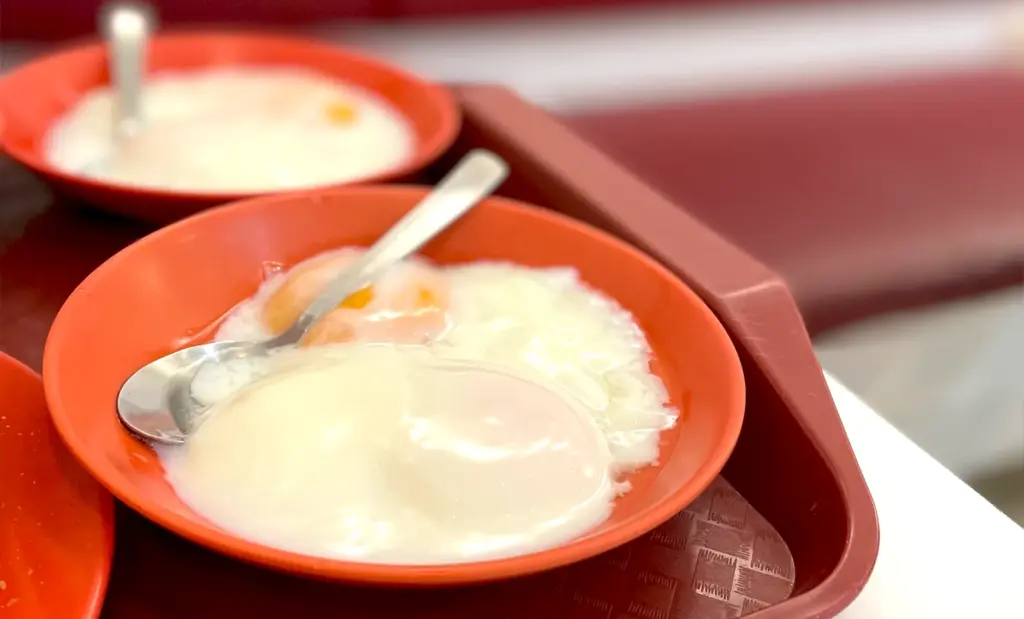 Freshly prepared soft-boiled eggs in a red bowl served with a spoon at a traditional Singapore breakfast spot.