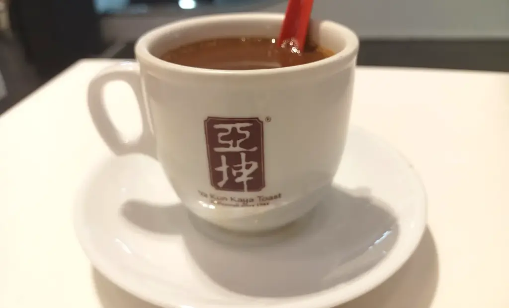 A ceramic cup of traditional Nanyang coffee stamped with the Ya Kun Kaya Toast logo and brand characters.
