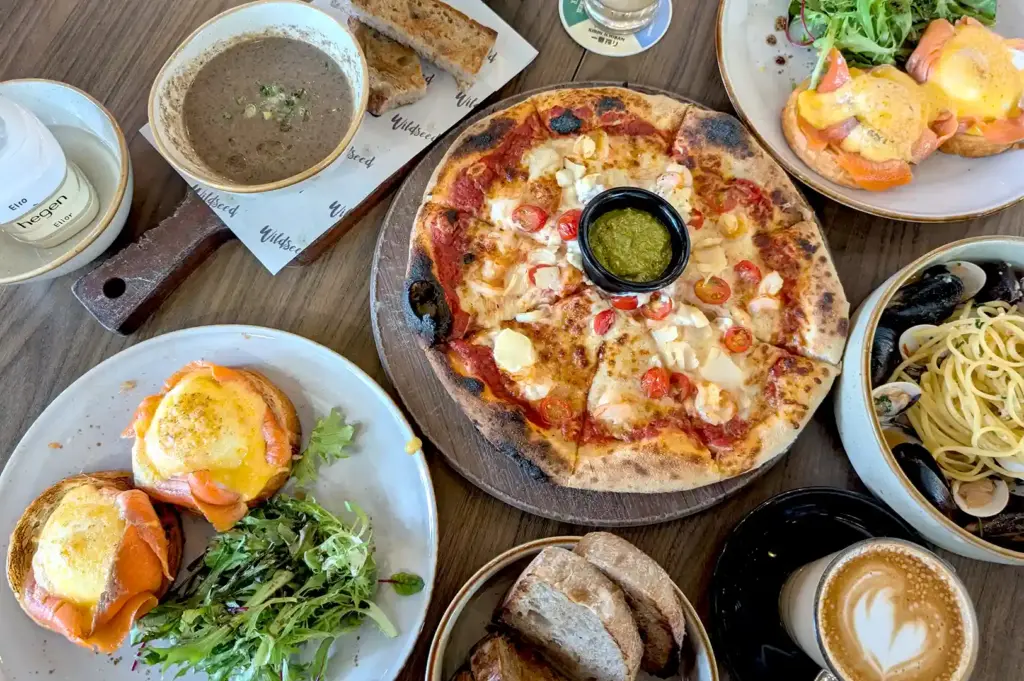 A top-down view of a hearty cafe spread featuring a wood-fired pizza with a dipping sauce bowl, eggs benedict with smoked salmon, mushroom soup, sliced bread, and pasta.