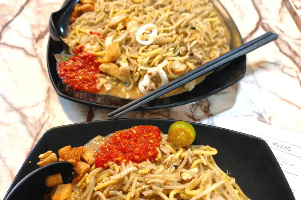 Black plates of signature Hokkien noodles garnished with sambal, lime, and seafood at Bao Er Cafe.