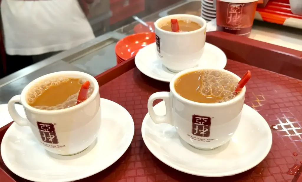 Freshly brewed cups of traditional Singaporean coffee with frothy foam served on saucers at a Ya Kun Kaya Toast counter.