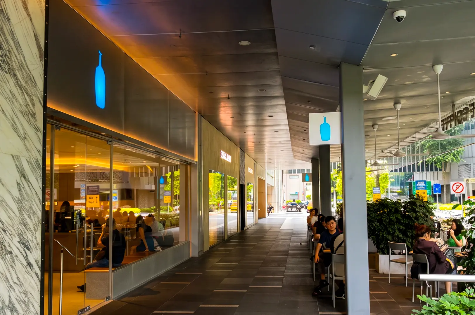 Blue Bottle Coffee exterior with its simple blue logo signage above the entrance and the interior blooming with warm yellow lights.