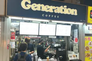 The storefront of Generation Coffee at Tekka Market, featuring a bold dark blue sign with gold lettering and a digital menu board displaying various drinks.