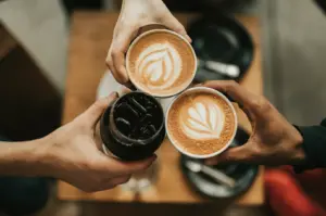 A high angle view of three coffee cups and glass held by three hands with latte art on two of the cups and a wooden table blurred in the background.