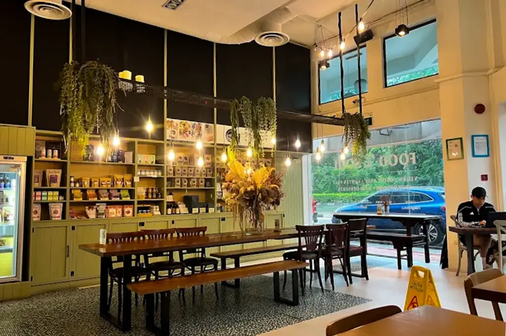 The Living Cafe interior with its design combined with hanging plants and warm yellow lights, a shelf at the back showing vegan products and a long wooden table in the center.