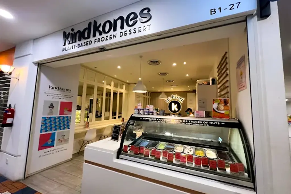 kind Kones exterior with a glass display showing available vegan ice cream flavors and their logo on top, the lightly illuminated interior shows white , clean tables and chairs.