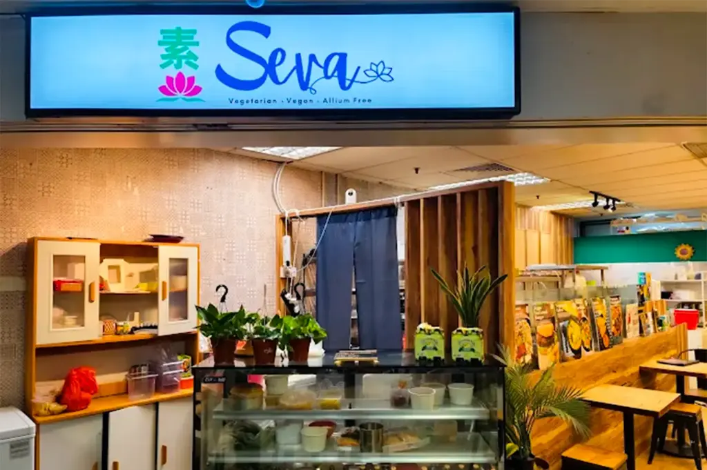 Seva Cafe exterior with its white lighted logo signage on top and the cozy interior visible inside with the counter and tables and chairs under warm lighitng.