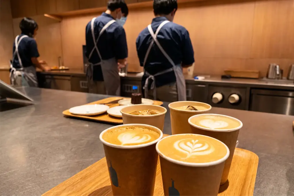 Five coffee cups arranged on a wooden tray on a counter with the staff working behind.
