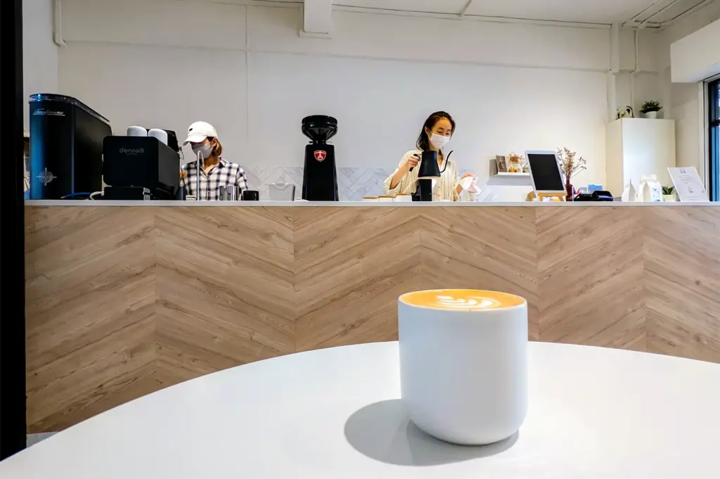 White coffee cup on a white table with the counter behind showing a staff working the counter at Zerah Coffee Roasters.