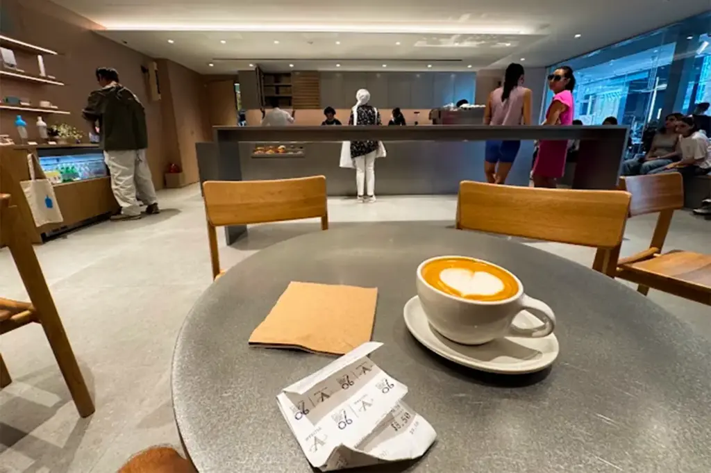An unoccupied table with a coffee served in a white mug and platter, the busy Blue Bottle Coffee interior in the background.