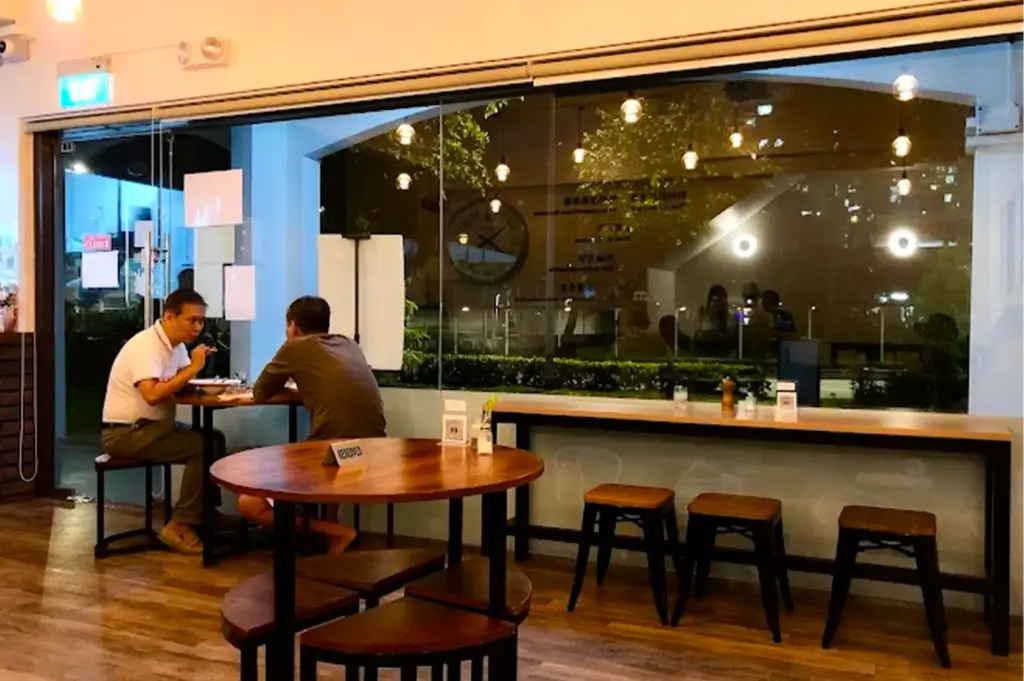 Loving Nature Fortunate Coffee Cafe SG interior with wooden and metal tables and chairs around and two dining customers seated near the glass entrance and window illuminated by warm yellow lights.