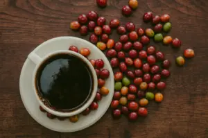 A top view of a white coffee cup with fresh coffee cherries scattered around on a wooden table.