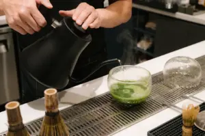 A freshly made matcha drink with how water being poured out of a pot in a glass cup on a counter.