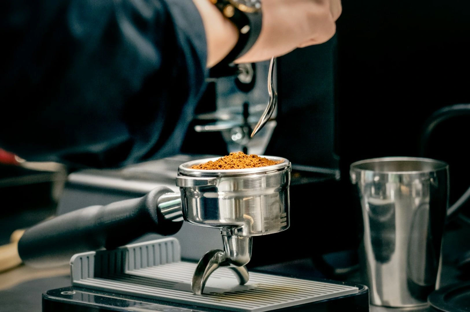 Close-up of a barista's hand holding a metal portafilter filled with freshly ground coffee. A coffee machine and metal cup are in the background.